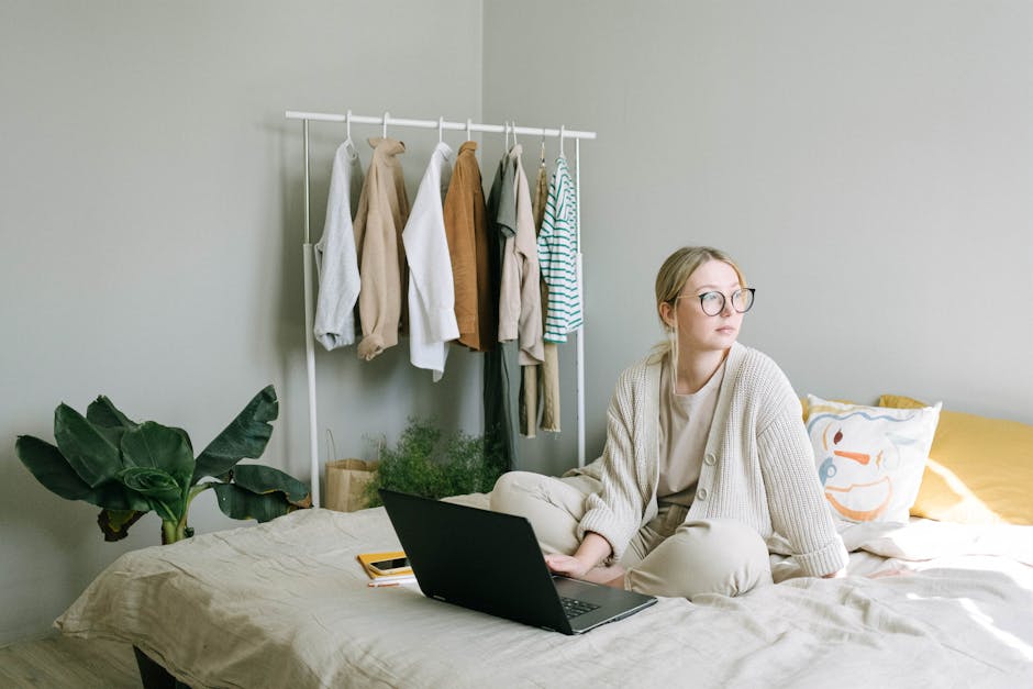Adult woman in eyeglasses working from home on a laptop, sitting on a bed with clothes rack nearby.