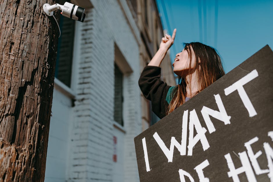 A woman gestures toward a CCTV camera while holding a protest sign in a city setting.